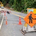 Road construction zone with orange traffic cones, barricades, and a workers-ahead warning sign on a rural road. A yellow construction vehicle and workers can be seen in the background, while a motorcyclist navigates through the partially blocked road. A black dog stands on the grassy area to the left.
