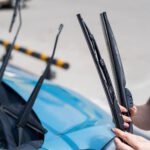 Person holding a new windshield wiper blade while standing next to a car with raised wiper arms, preparing to replace the old blades.
