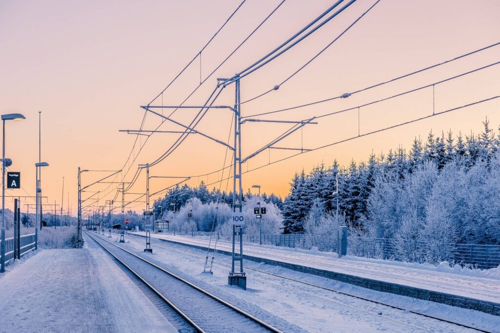 Snow-covered train station platform in Sweden at sunset, with electrical power lines overhead and frost-covered pine trees in the background creating a serene winter landscape.
