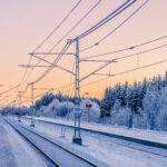 Snow-covered train station platform in Sweden at sunset, with electrical power lines overhead and frost-covered pine trees in the background creating a serene winter landscape.