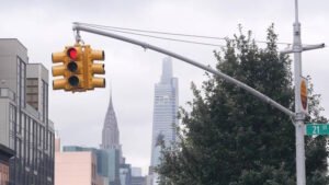 A yellow traffic light shows a red signal at an intersection on 21st Street. The traffic light is mounted on a long horizontal pole extending from a streetlight. In the background, iconic New York City skyscrapers, including the Chrysler Building and a modern glass tower, rise into a cloudy sky. The scene includes urban buildings, trees, and overhead wires.