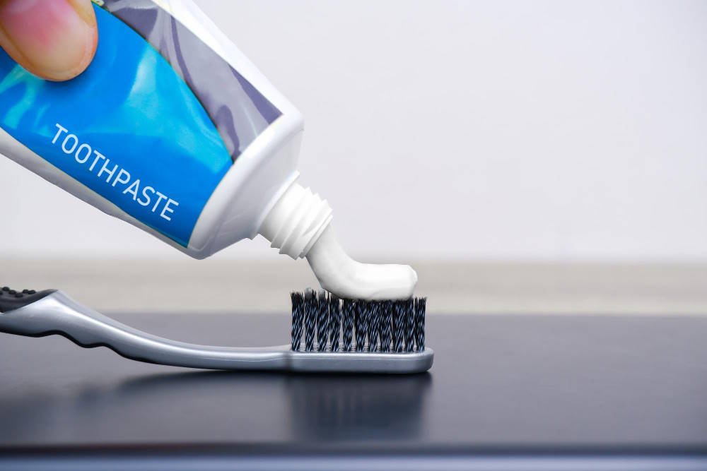 Close-up of a person squeezing white toothpaste from a blue tube labeled "TOOTHPASTE" onto a toothbrush against a neutral background, demonstrating the first step in the DIY headlight cleaning process.