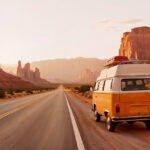 A yellow and white VW Vanagon van on an empty desert highway surrounded by red rock formations in Monument Valley during golden hour sunset.