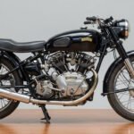 Side view of a classic Vincent Black Shadow motorcycle featuring a black fuel tank with gold logo, exposed V-twin engine, chrome exhaust, and a vintage registration plate "VV 459" on the front fender.