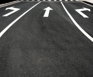 A clean asphalt roadway featuring three marked lanes, each with bold white directional arrows. The left lane has a left-turn arrow, the center lane shows a straight arrow, and the right lane has a right-turn arrow. A pedestrian crosswalk is visible in the background, indicating an upcoming intersection or crossing zone. 
