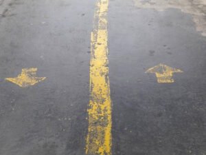 A worn asphalt surface showing yellow-painted traffic markings. A solid yellow line runs vertically through the center, with two directional arrows on either side—one pointing left and the other pointing right—indicating bidirectional flow or lane separation. The markings appear faded, suggesting heavy use over time.