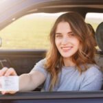 Smiling young woman sitting in a car holding out her driver's license through the window with a blurred green field in the background.
