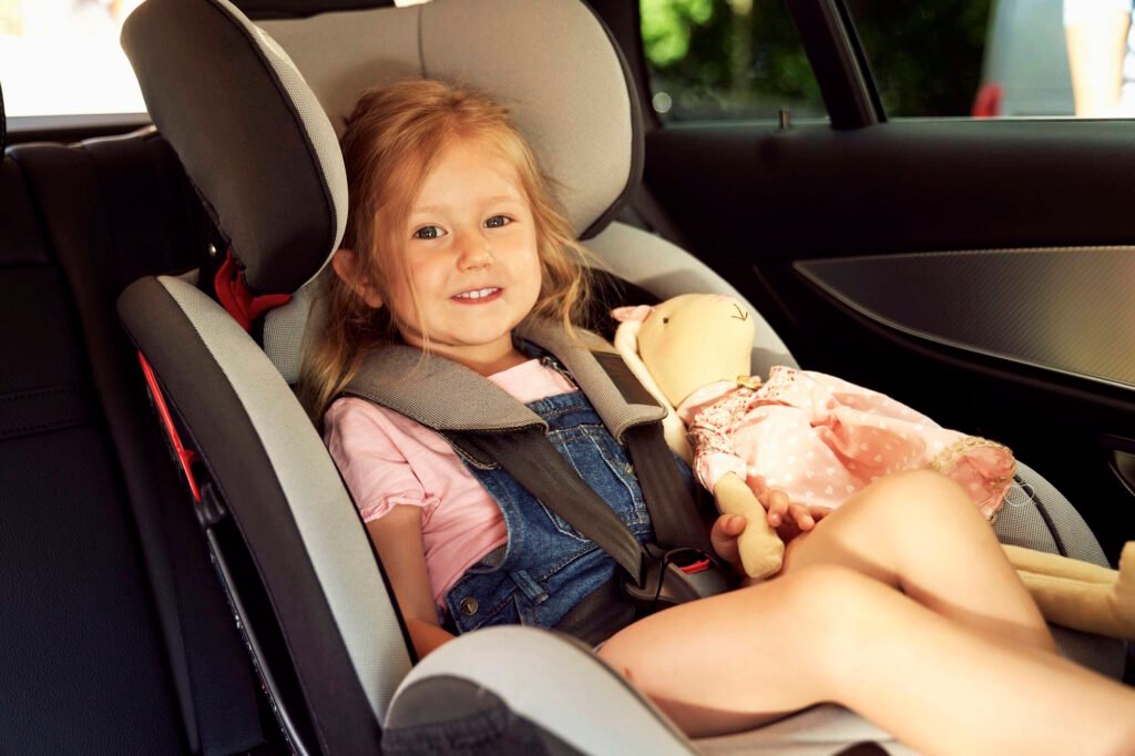 Young girl with blonde hair sitting safely in a forward-facing car seat with five-point harness, holding a doll, demonstrating proper child passenger safety in a vehicle's back seat.RetryClaude can make mistakes. Please double-check responses.