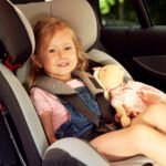 Young girl with blonde hair sitting safely in a forward-facing car seat with five-point harness, holding a doll, demonstrating proper child passenger safety in a vehicle's back seat.RetryClaude can make mistakes. Please double-check responses.