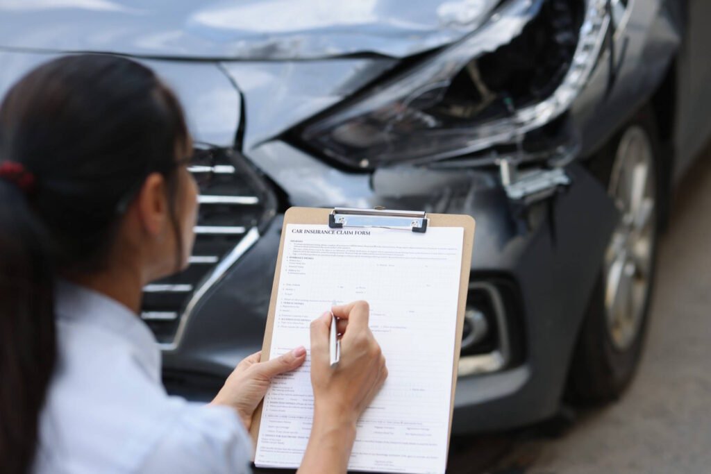 Insurance adjuster filling out car insurance claim form on clipboard while examining damaged vehicle after accident.