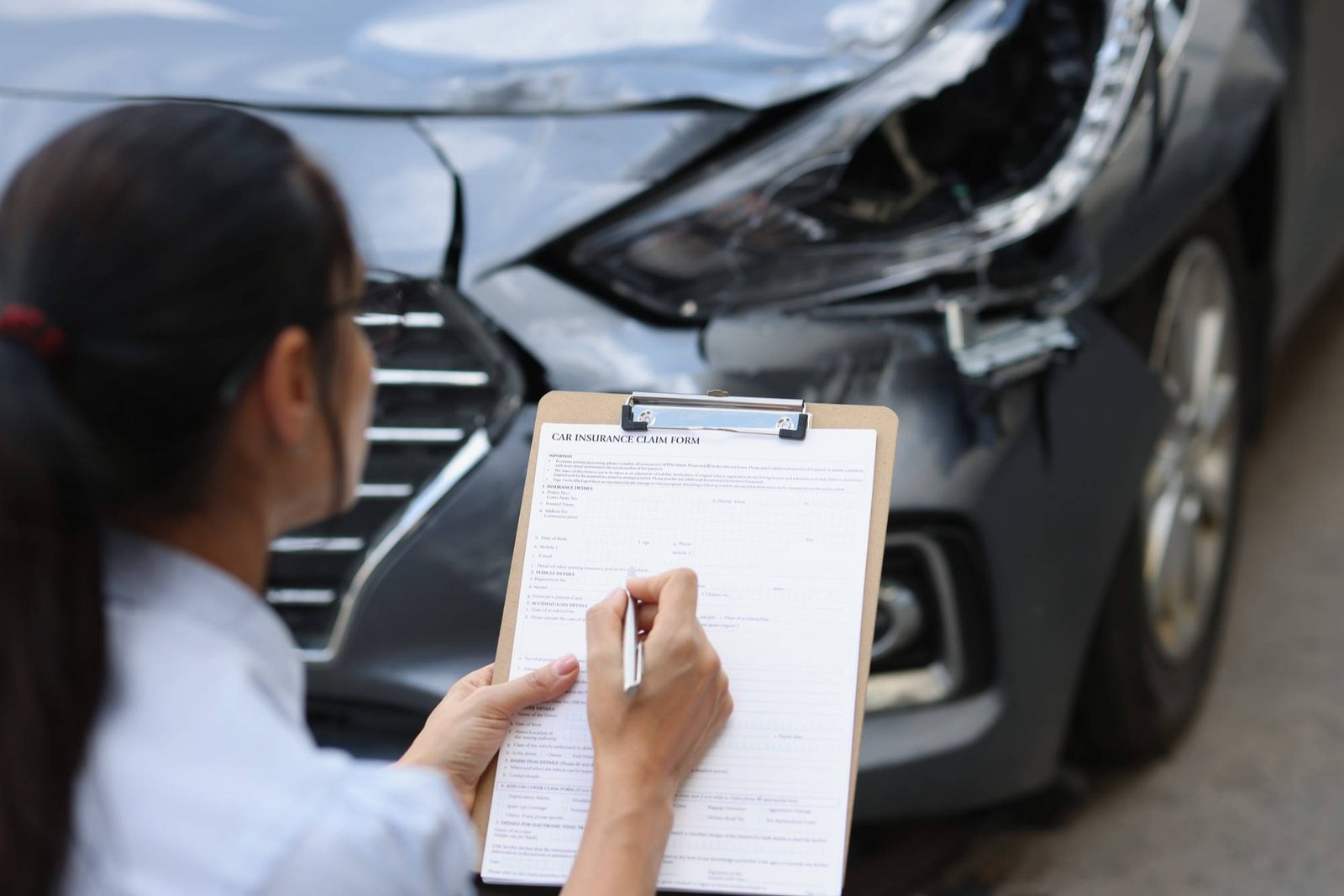 Insurance adjuster filling out car insurance claim form on clipboard while examining damaged vehicle after accident.