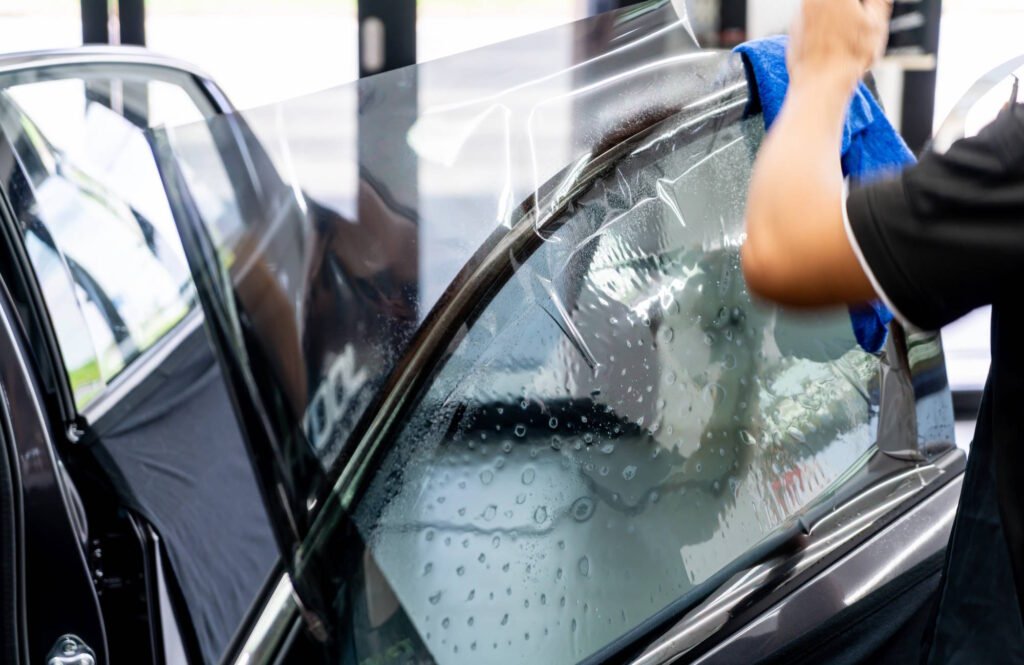 A technician's hand using a blue cloth to apply clear protective film to a wet car window during the tinting installation process, with water droplets visible on the glass surface.