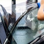 A technician's hand using a blue cloth to apply clear protective film to a wet car window during the tinting installation process, with water droplets visible on the glass surface.