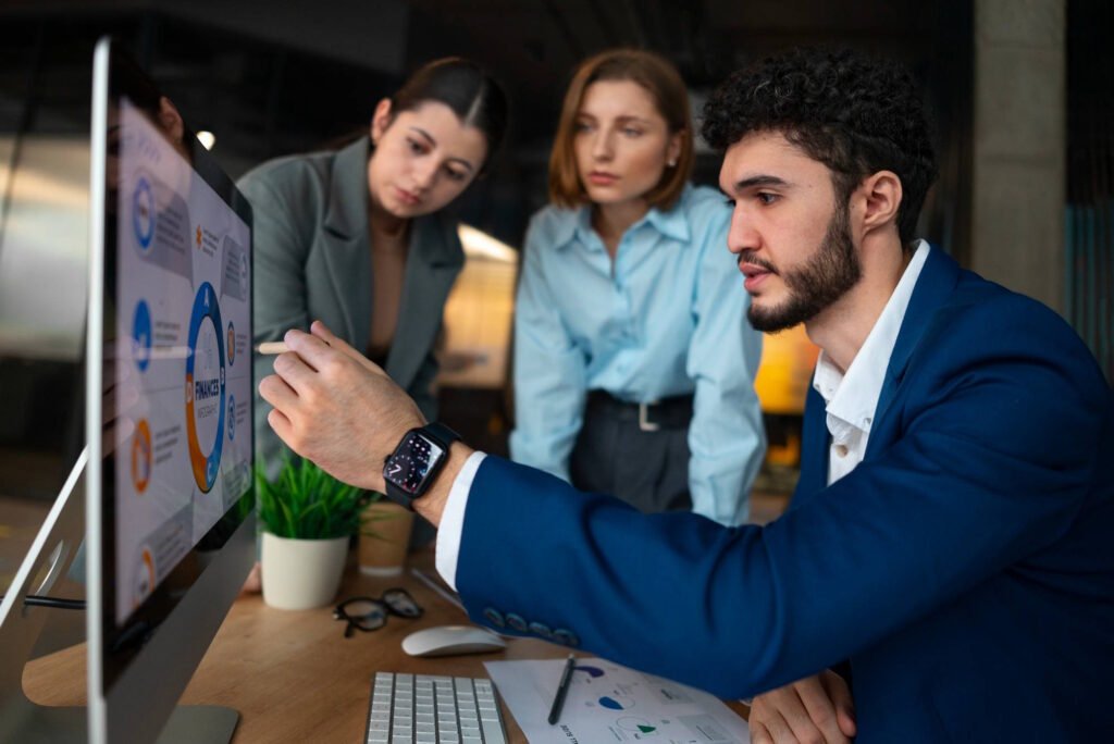 Three business professionals analyzing data on a computer screen - a man in a blue suit pointing at charts while two women colleagues observe and collaborate in a modern office setting.
