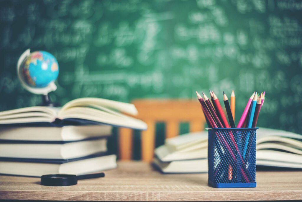 Classroom desk with stacked books, colored pencils in blue mesh holder, small globe, and green chalkboard with math formulas in background.