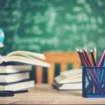 Classroom desk with stacked books, colored pencils in blue mesh holder, small globe, and green chalkboard with math formulas in background.
