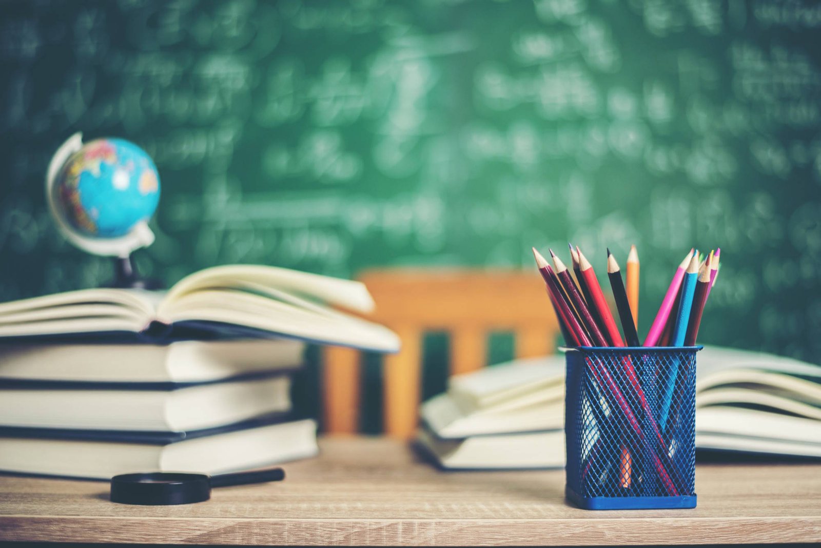 Classroom desk with stacked books, colored pencils in blue mesh holder, small globe, and green chalkboard with math formulas in background.