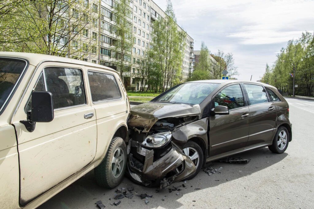 Two damaged cars after a collision on a residential street, showing a beige older vehicle and a black sedan with significant front-end damage, debris scattered on the road.