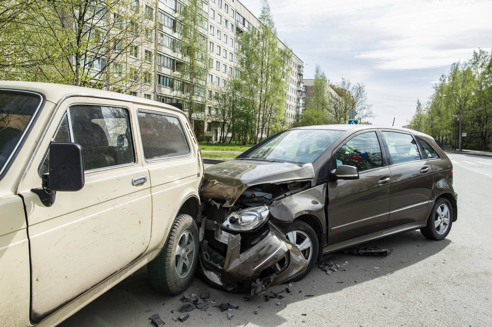 Two damaged cars after a collision on a residential street, showing a beige older vehicle and a black sedan with significant front-end damage, debris scattered on the road.
