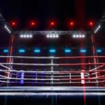 Professional wrestling ring with dramatic red and blue lighting in a large arena, featuring multiple rope levels and professional broadcast lighting setup