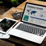MacBook laptop displaying a business analytics dashboard titled "Company's Growth" with world map, 85% stock market indicator, and financial data tables, alongside a tablet showing 08:15, glasses, notebook, and coffee cup on wooden desk