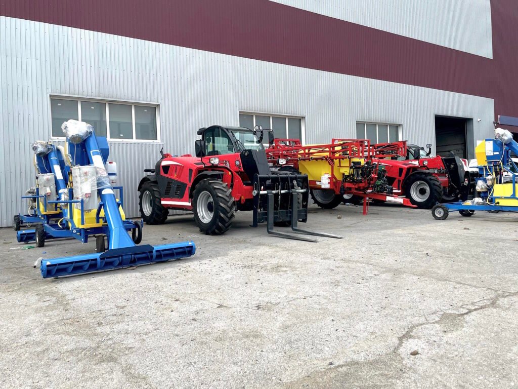 Red telehandler with forks parked next to blue and yellow agricultural equipment outside gray metal warehouse building