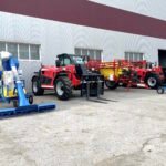 Red telehandler with forks parked next to blue and yellow agricultural equipment outside gray metal warehouse building