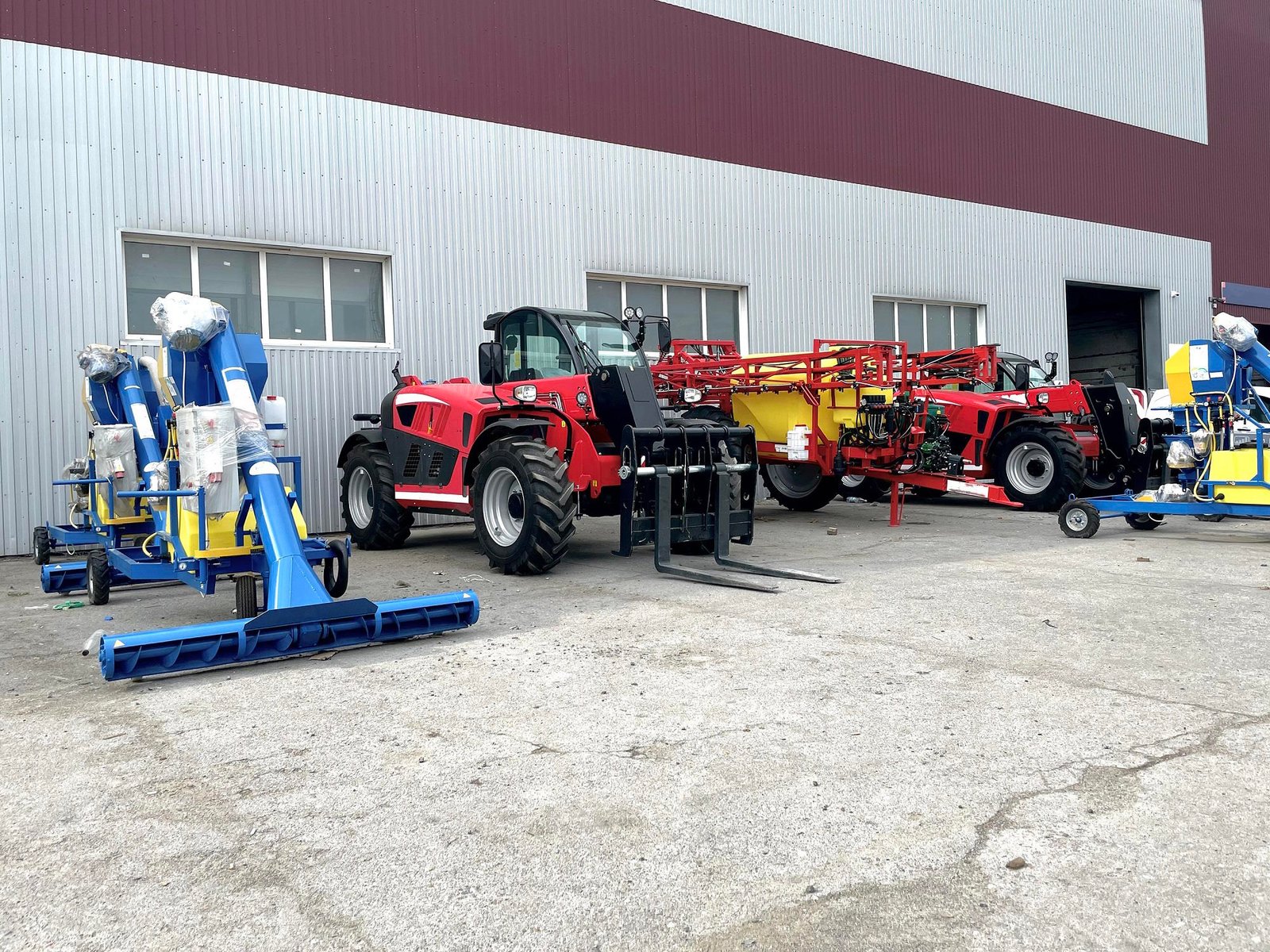Red telehandler with forks parked next to blue and yellow agricultural equipment outside gray metal warehouse building