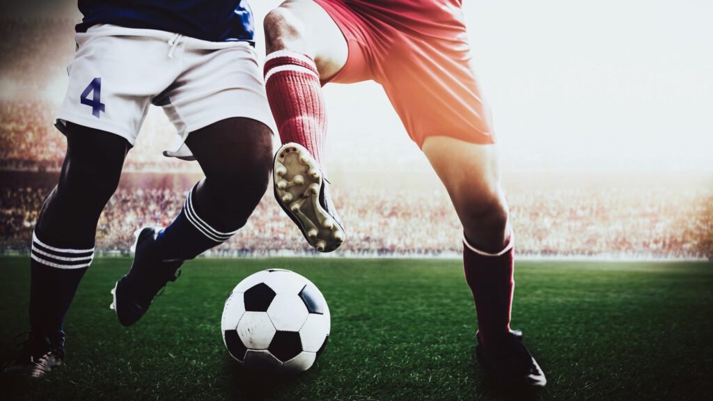 Two football players in action competing for the ball on a grass pitch with stadium crowd in background.