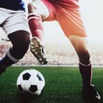 Two football players in action competing for the ball on a grass pitch with stadium crowd in background.