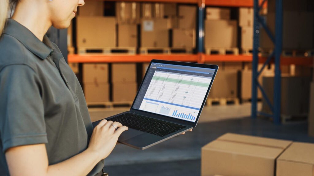 Warehouse worker using laptop computer to manage inventory data and analytics in modern distribution center with storage shelves.