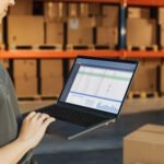 Warehouse worker using laptop computer to manage inventory data and analytics in modern distribution center with storage shelves.