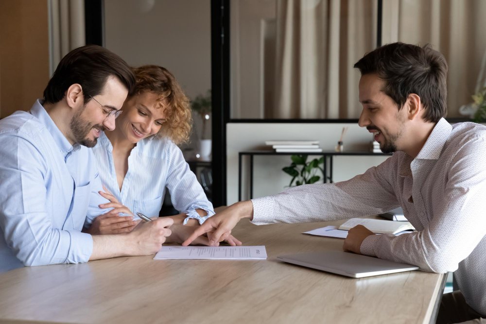 Three business professionals sitting at a wooden conference table reviewing documents together, with a man and woman on the left side consulting with another man across the table in a modern office setting.
