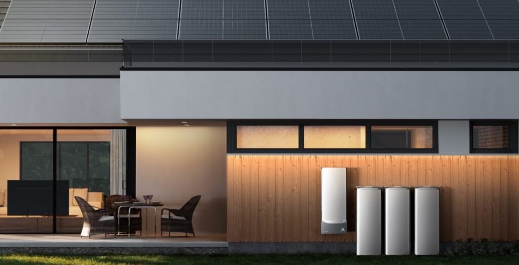 Modern home exterior at dusk with solar panels on the roof and four white cylindrical EcoFlow OCEAN Pro battery units installed against a wood-paneled wall, featuring large glass windows showing warm interior lighting and contemporary furniture.