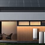 Modern home exterior at dusk with solar panels on the roof and four white cylindrical EcoFlow OCEAN Pro battery units installed against a wood-paneled wall, featuring large glass windows showing warm interior lighting and contemporary furniture.