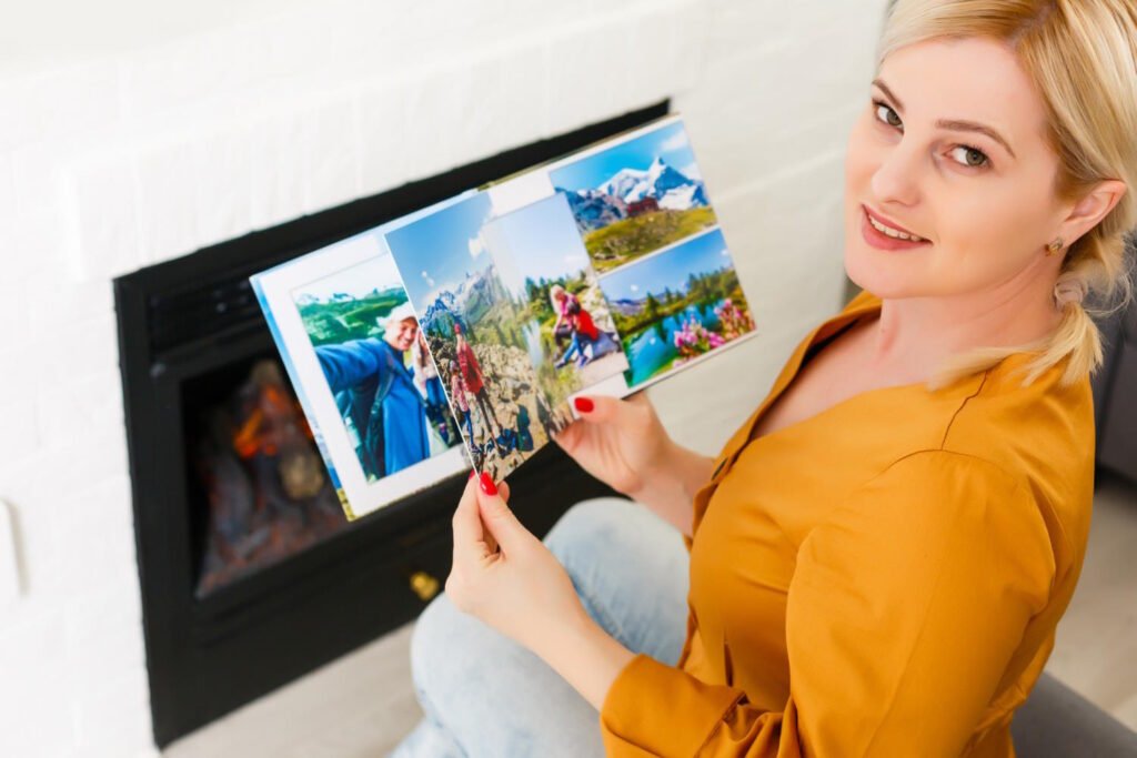 Woman in yellow shirt smiling while holding printed travel photos in front of computer screen displaying vacation images.