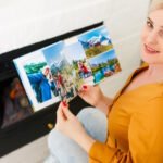 Woman in yellow shirt smiling while holding printed travel photos in front of computer screen displaying vacation images.