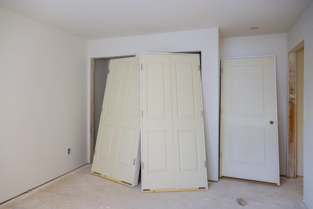 Several white interior doors leaning against a wall in an unfinished room during construction or renovation, with one door partially installed in a doorway frame.