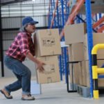 Warehouse worker in a red plaid shirt and cap lifting stacked cardboard boxes manually, demonstrating the challenges of heavy load handling without equipment.