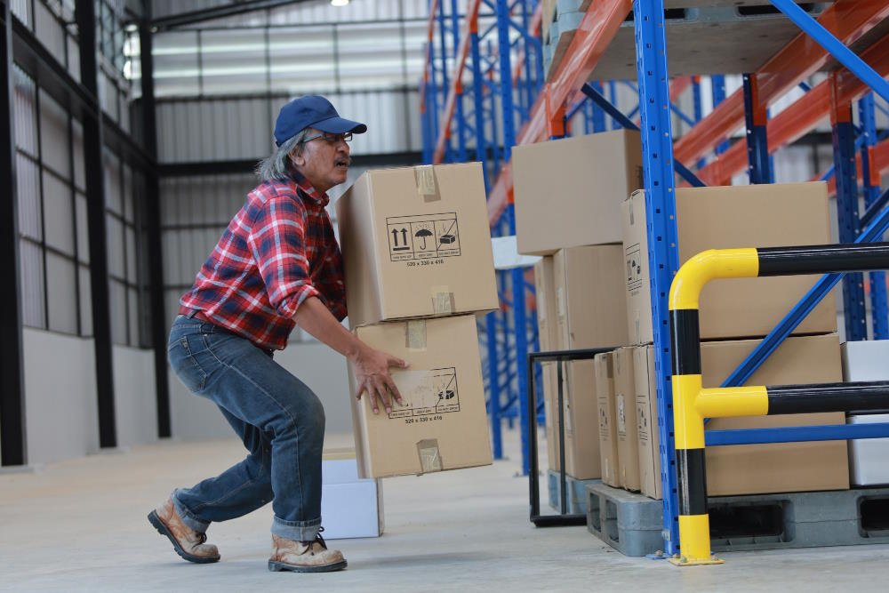Warehouse worker in a red plaid shirt and cap lifting stacked cardboard boxes manually, demonstrating the challenges of heavy load handling without equipment.