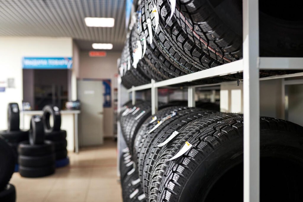 Interior view of a modern tire shop showing organized tire storage with black tires displayed on white metal shelving racks, with additional tires and automotive service area visible in the background