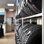Interior view of a modern tire shop showing organized tire storage with black tires displayed on white metal shelving racks, with additional tires and automotive service area visible in the background