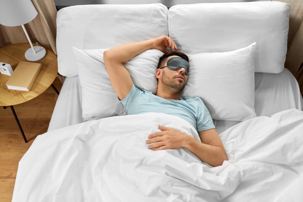 Man sleeping peacefully in white bedding wearing a sleep mask, lying on his back in a modern bedroom with wooden nightstand and lamp
