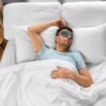 Man sleeping peacefully in white bedding wearing a sleep mask, lying on his back in a modern bedroom with wooden nightstand and lamp