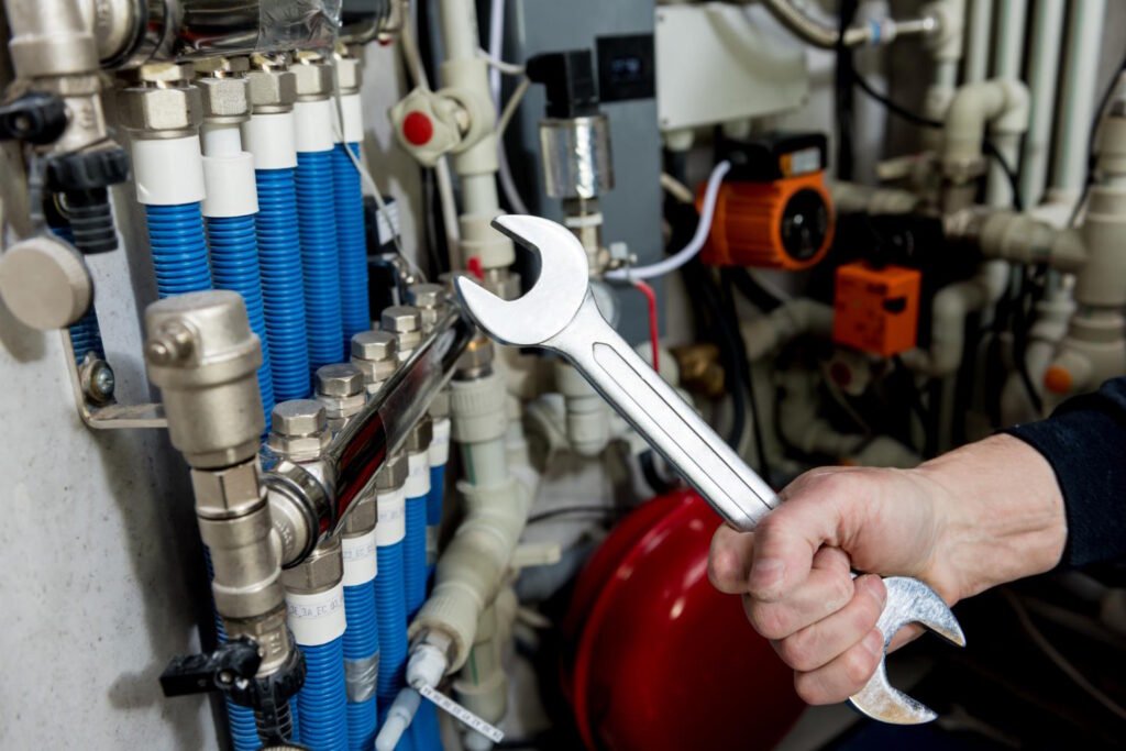 Hand holding a wrench near industrial heating system components including blue coiled tubes, metal fittings, and control valves during maintenance work