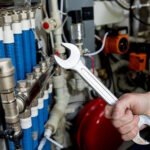 Hand holding a wrench near industrial heating system components including blue coiled tubes, metal fittings, and control valves during maintenance work