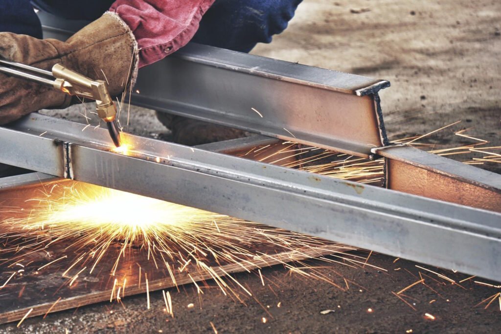 A skilled welder in protective gloves operates a cutting torch on steel beams in an industrial fabrication shop, creating bright sparks and demonstrating precision metalworking techniques used in custom metal forming and manufacturing processes.