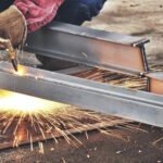 A skilled welder in protective gloves operates a cutting torch on steel beams in an industrial fabrication shop, creating bright sparks and demonstrating precision metalworking techniques used in custom metal forming and manufacturing processes.