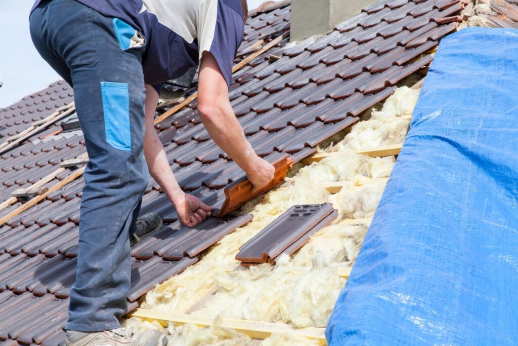 Roofer installing clay tiles on residential roof with exposed insulation and blue tarp visible during roofing project