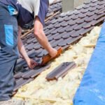 Roofer installing clay tiles on residential roof with exposed insulation and blue tarp visible during roofing project
